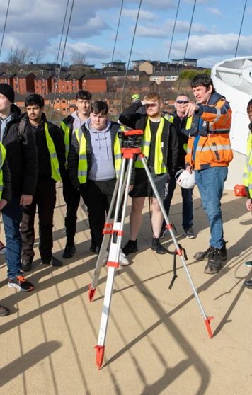 A group of students and lecturers in high-visibility vests gathered around a surveying instrument on the Stockingfield Bridge, listening to an instructor providing guidance. A group of students and lecturers in high-visibility vests gathered around a surveying instrument on the Stockingfield Bridge, listening to an instructor providing guidance.